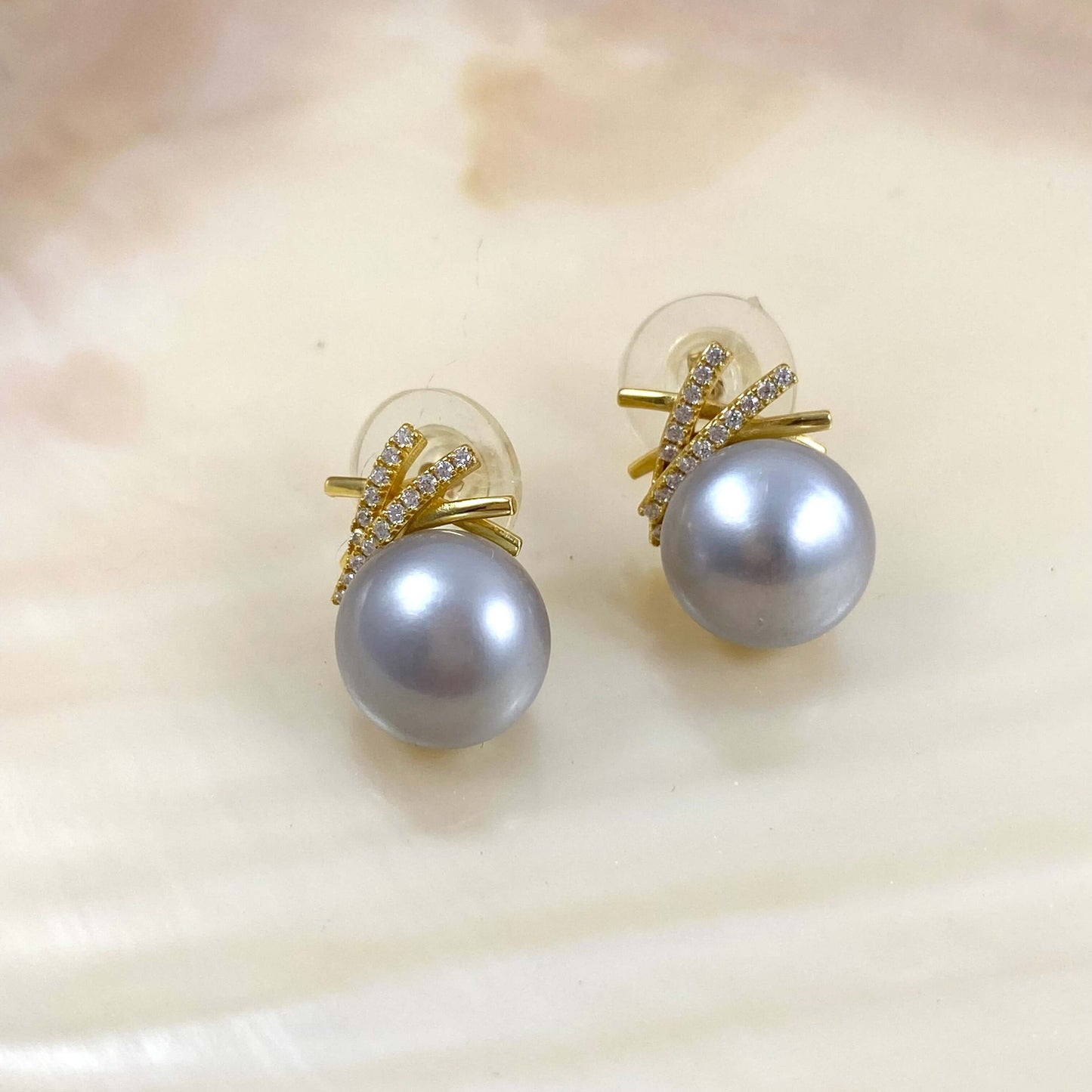A pair of gold stud earrings featuring a large gray pearl and a small gold branch-shaped setting accented with tiny clear stones, displayed on a white clam shell.