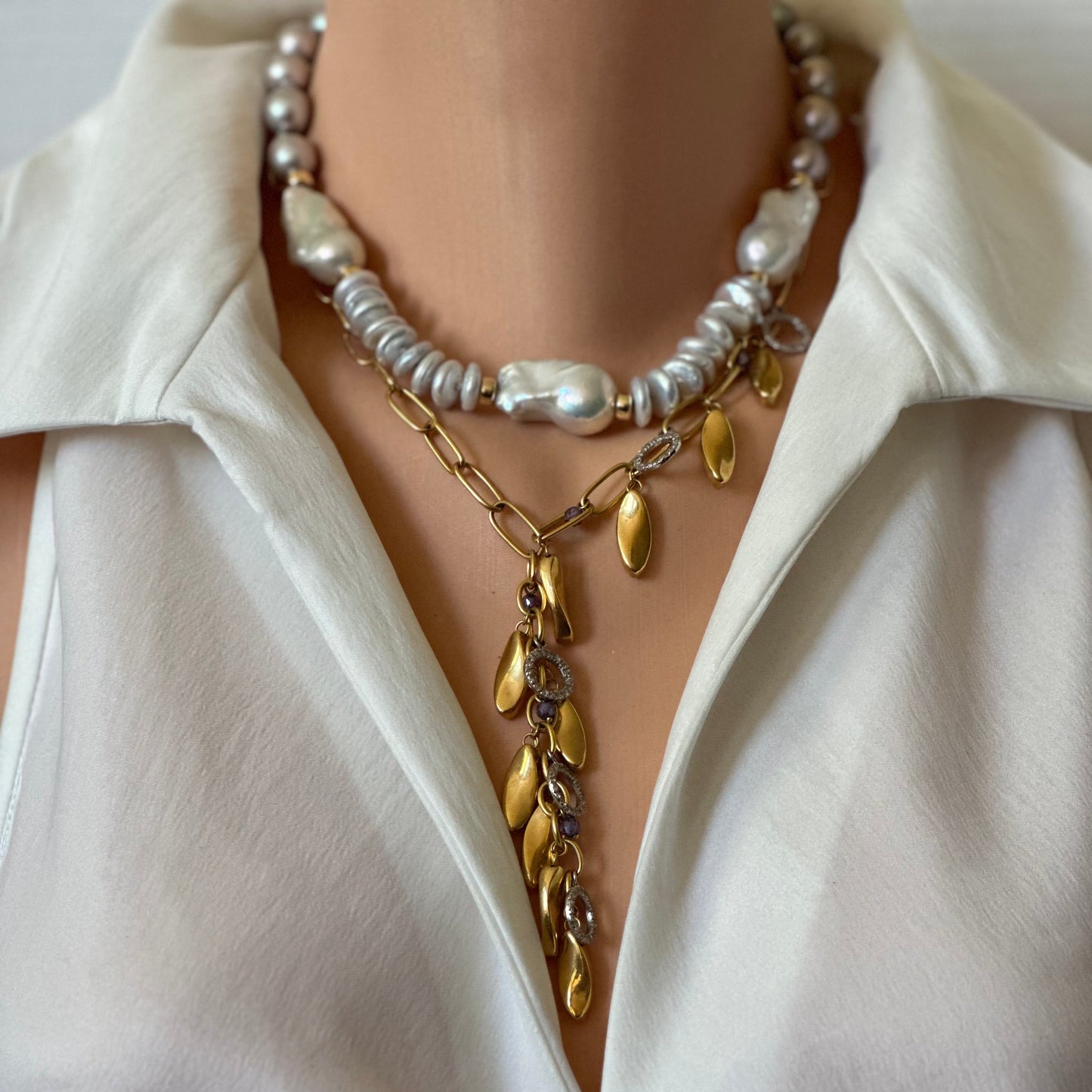 A close-up of a person wearing a layered necklace set over a white collared blouse. The necklace features a top layer with white and silver baroque pearls, a middle layer with a gold chain and gold leaf-shaped charms, and a bottom layer with a gold chain and gold leaf charms.
