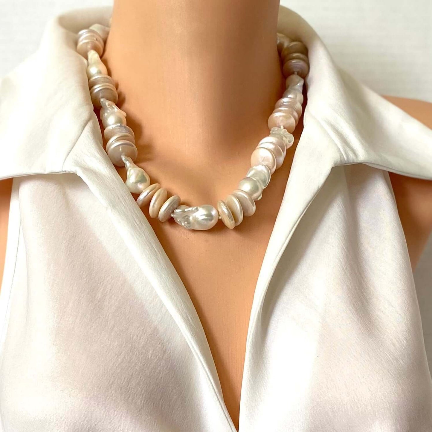 A close-up of a baroque pearl necklace worn on a mannequin or model torso, styled with a white satin collar blouse against a light neutral background.