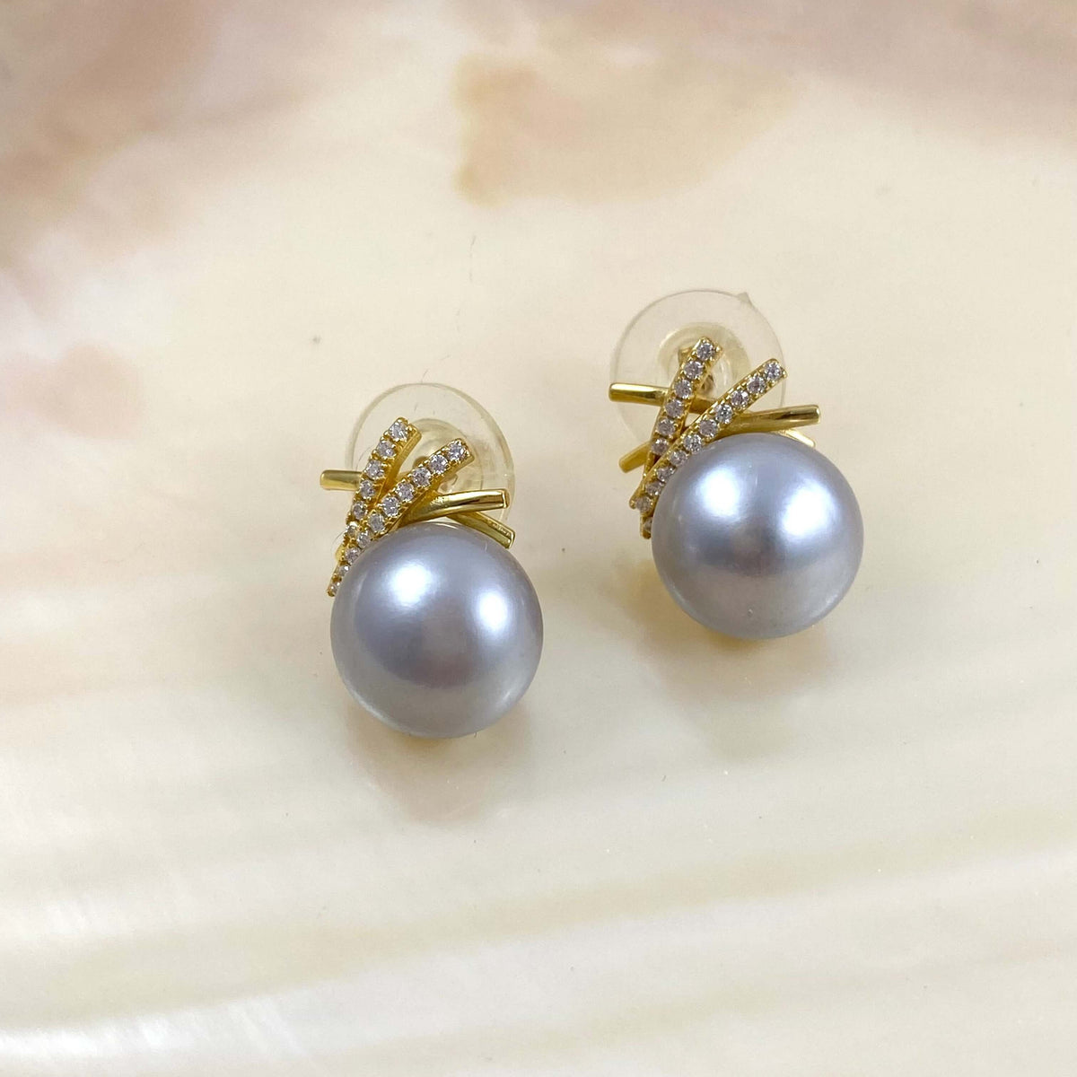 A pair of gold stud earrings featuring a large gray pearl and a small gold branch-shaped setting accented with tiny clear stones, displayed on a white clam shell.