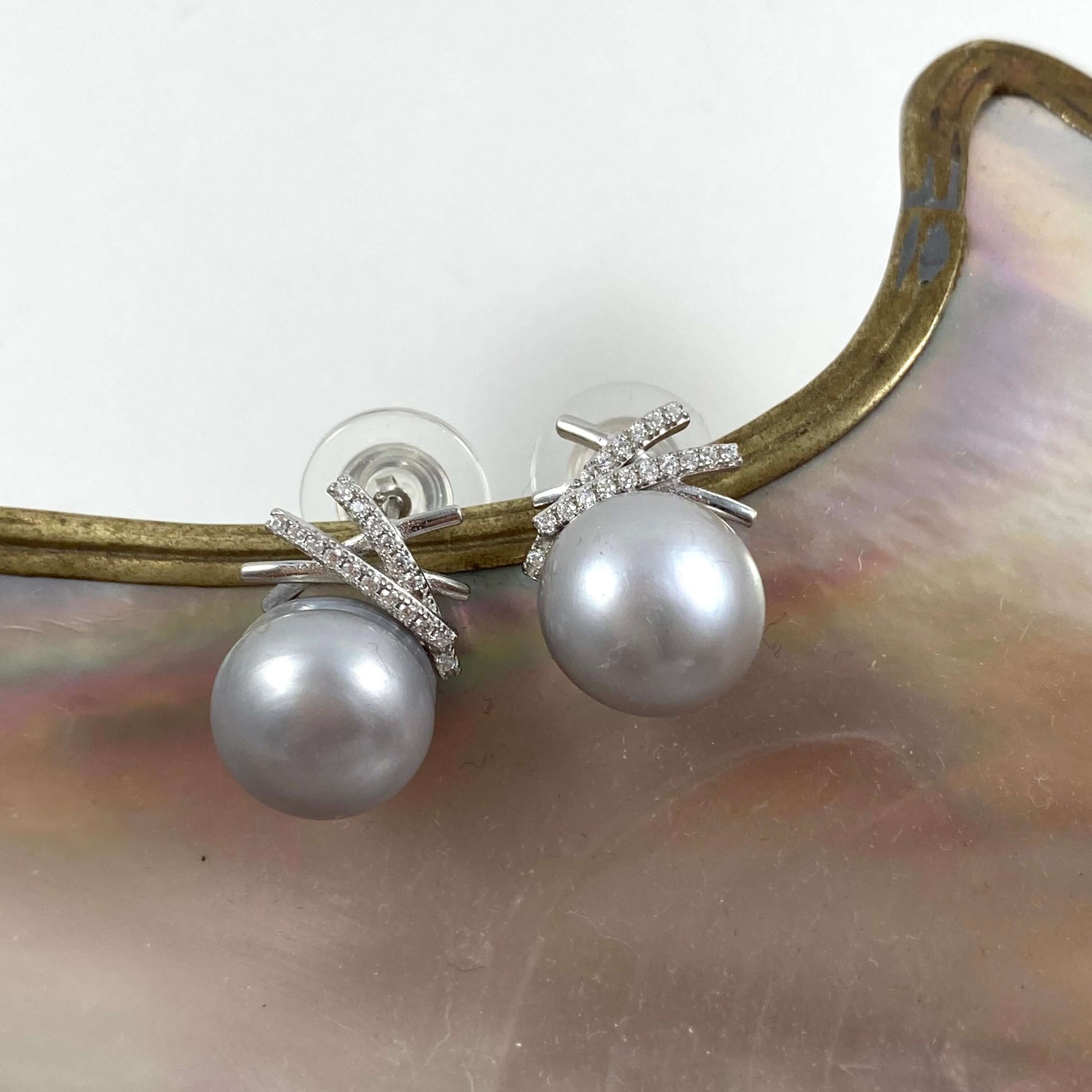 A pair of pearl stud earrings with silver-toned cross-shaped settings accented by small clear stones, resting on a curved iridescent shell piece against a white background.