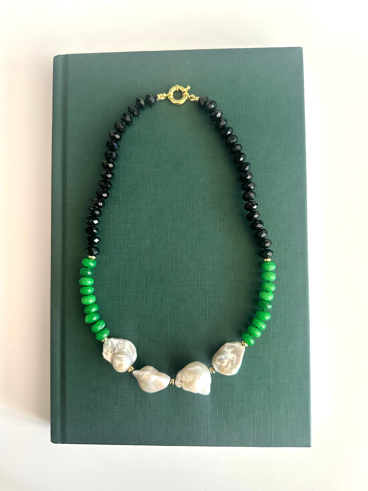 A beaded necklace with black faceted beads, bright green beads, and 4 irregular white baroque pearls, displayed on a dark green book against a white background.