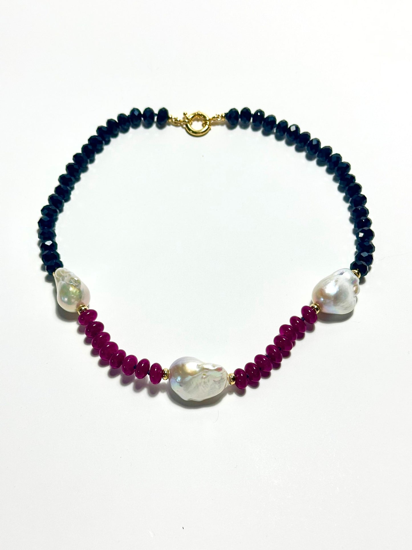 A beaded necklace featuring BLACK  faceted beads, deep magenta red beads, and three large white baroque pearls, finished with a gold clasp, shown on a plain white background.