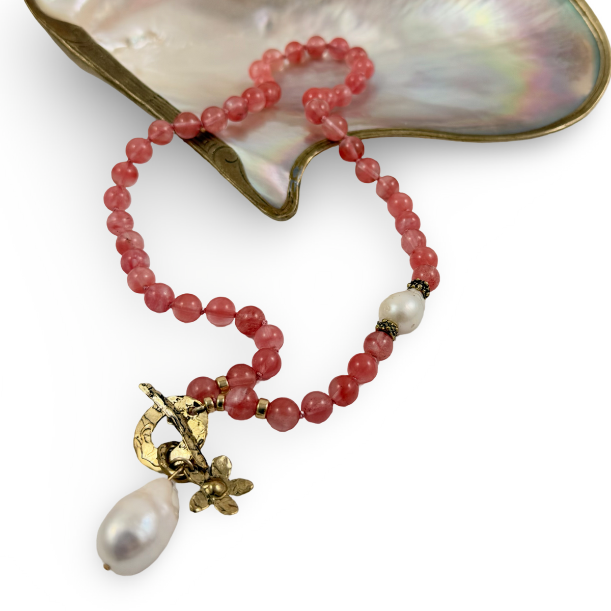 A beaded necklace with pinkish-red faceted beads, a white pearl accent, and a gold-tone floral pendant with a teardrop pearl, displayed on a white background with a large iridescent shell prop.