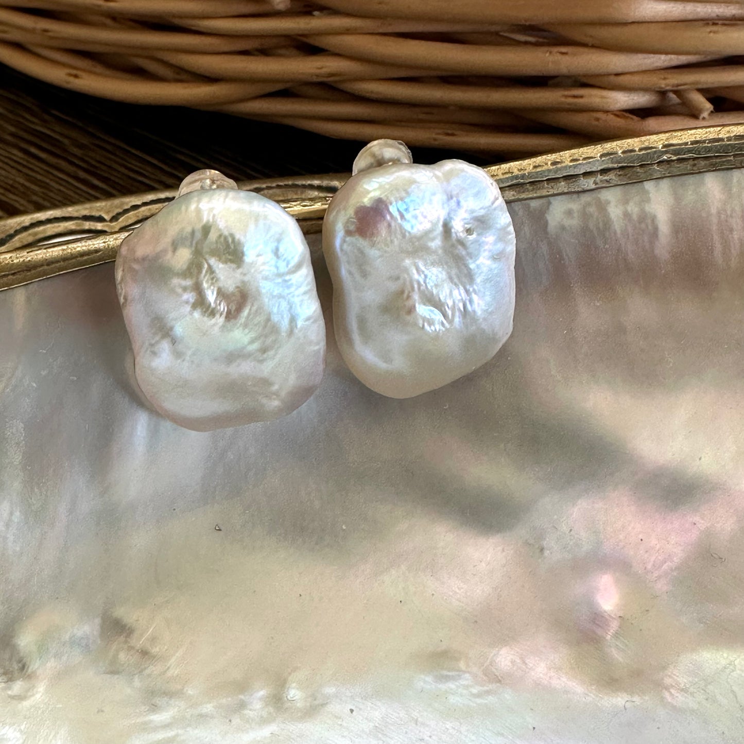 White rectangular baroque pearl stud earrings hanging from shell with wicker basket backdrop, showing radiant luster and elegant design.