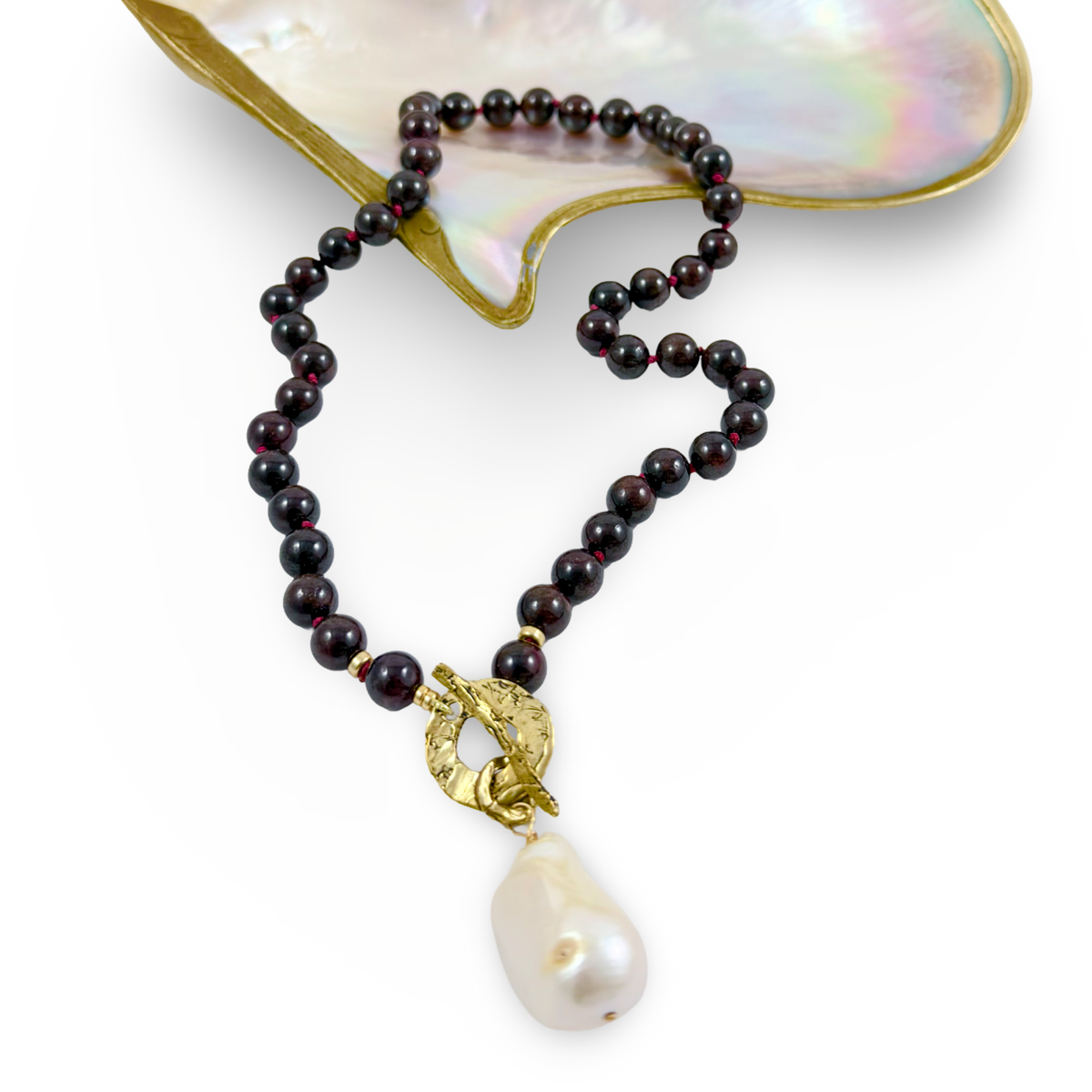A beaded necklace with dark red beads, a gold-toned toggle clasp, and a large white baroque pearl pendant, displayed on a white background with a mother-of-pearl shell prop.