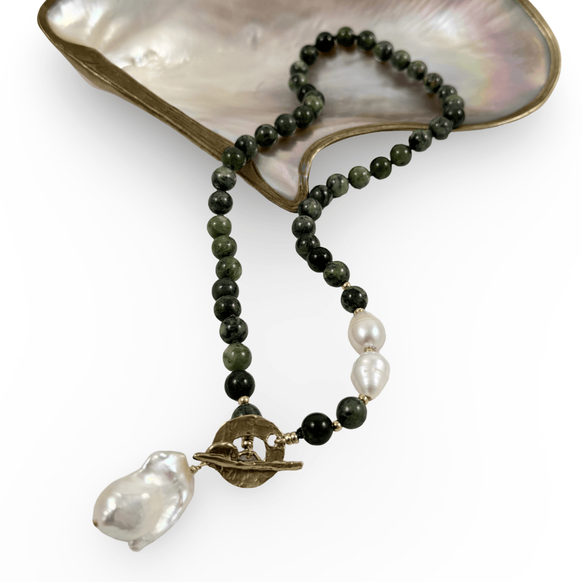 A beaded necklace with dark green and black round beads, white baroque pearls, and a gold toggle clasp, displayed on a white background with a mother-of-pearl shell prop.