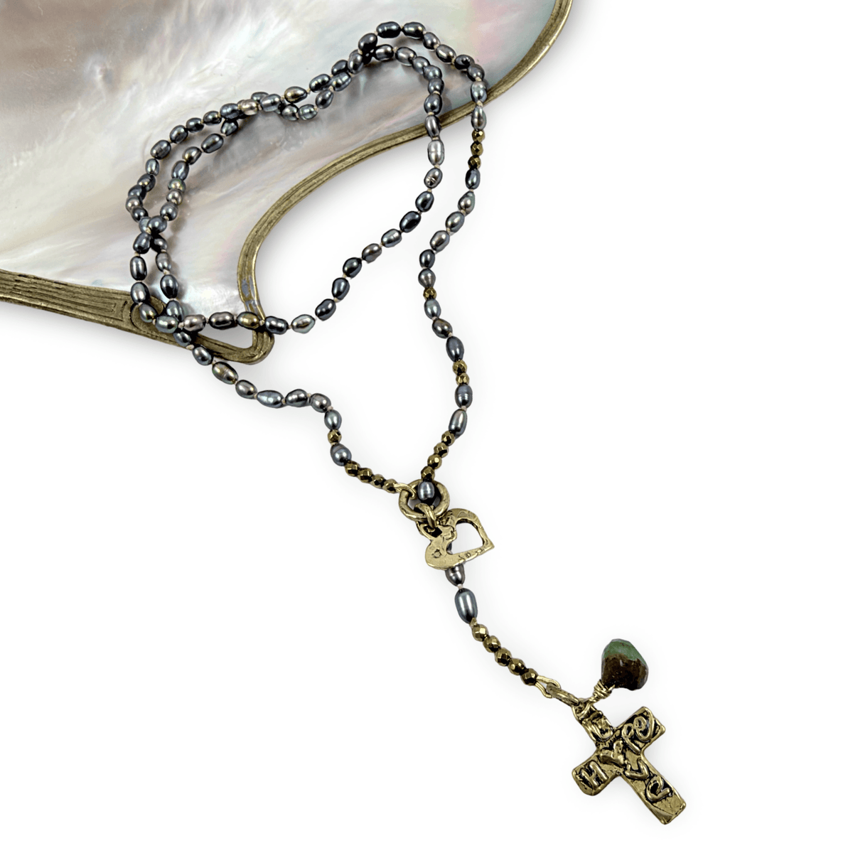A beaded rosary necklace with dark gray and black pearls, antique bronze-toned metal links, a small heart-shaped charm, and a cross pendant, displayed on a white background with a curved iridescent shell prop.