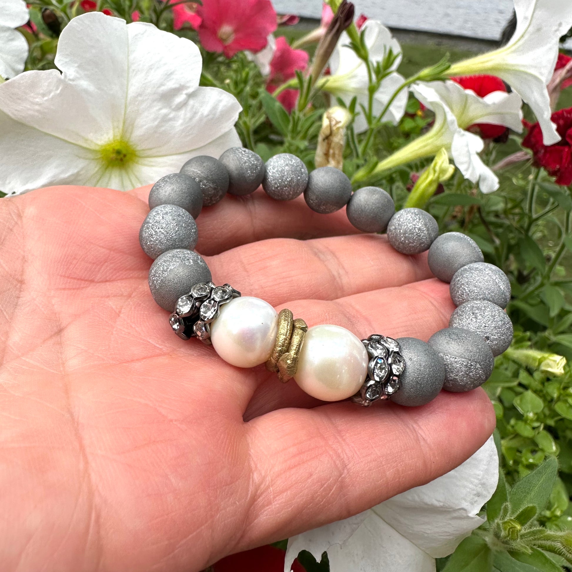 Close-up of grey druzy agate and pearl bracelet held outdoors near foliage