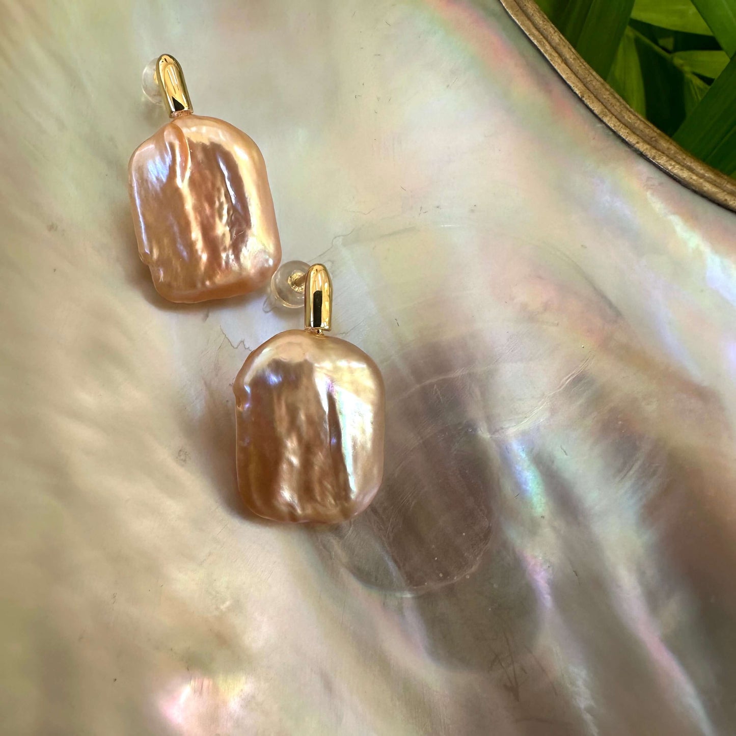 Golden peach pearl drop earrings photographed on white shell with green leaves, showing natural shimmer and refined gold finish.
