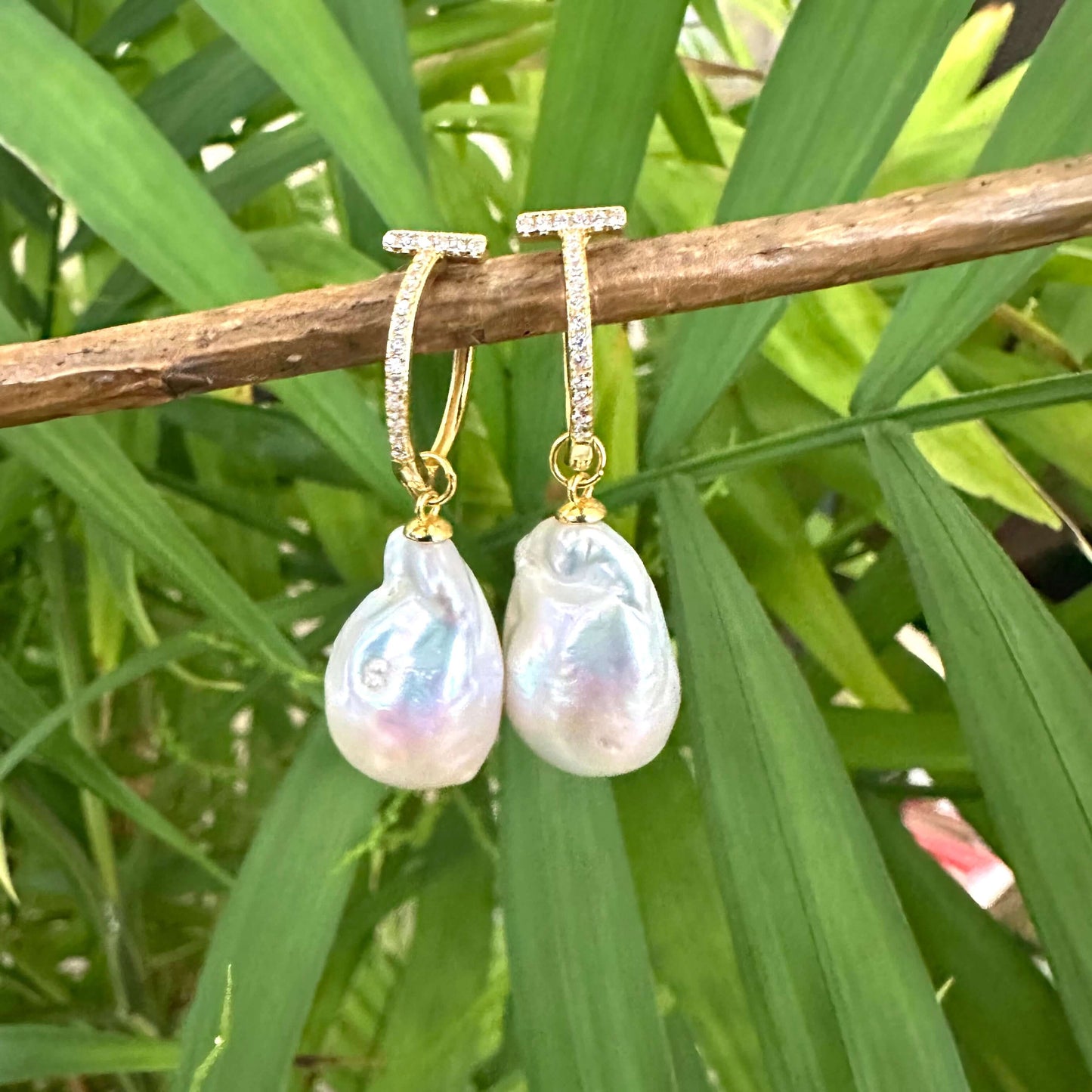 White Baroque Pearl Earrings – gold version held by hand against green leaf backdrop