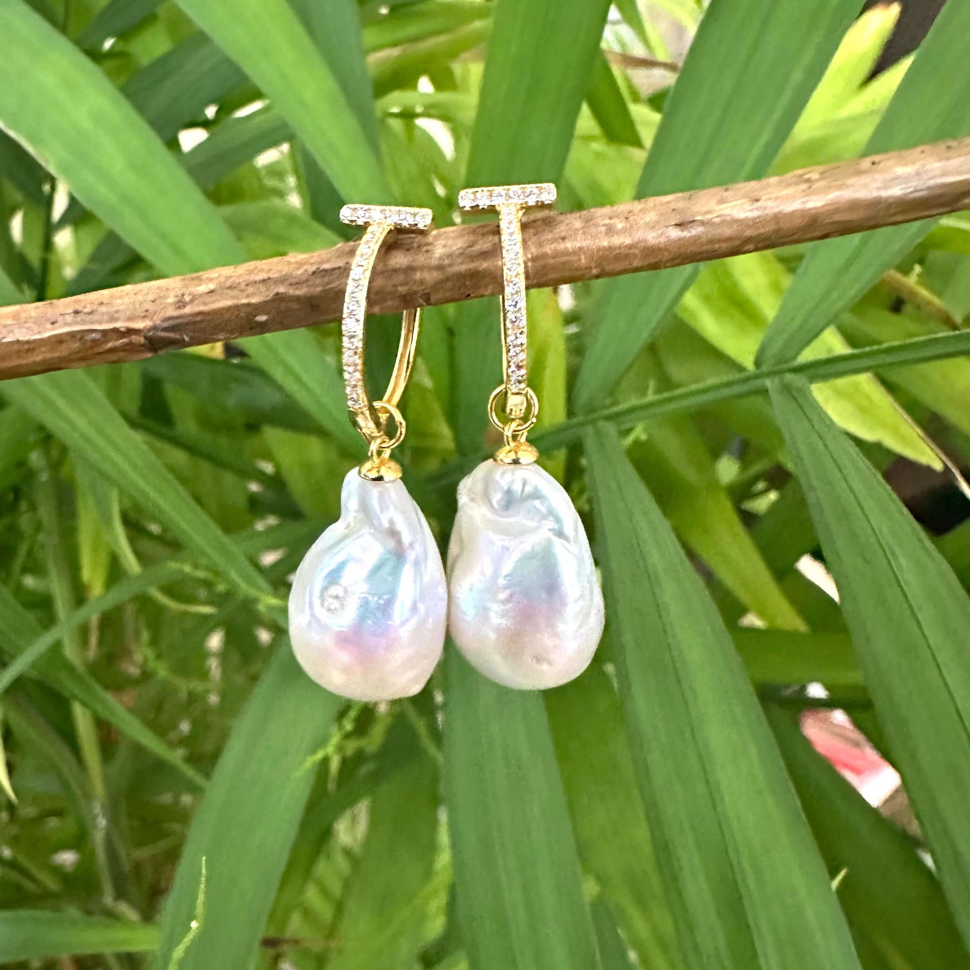 White Baroque Pearl Earrings – gold version held by hand against green leaf backdrop