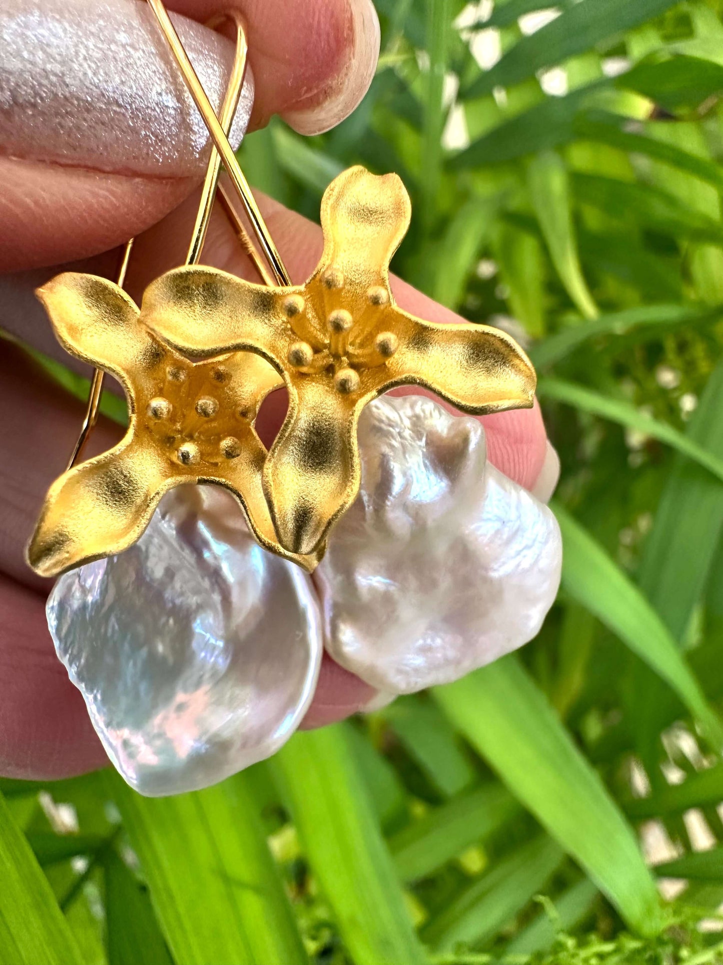 Keshi Pearl Earrings with white petal pearls and gold vermeil flower hooks held in hand against green leaf background.