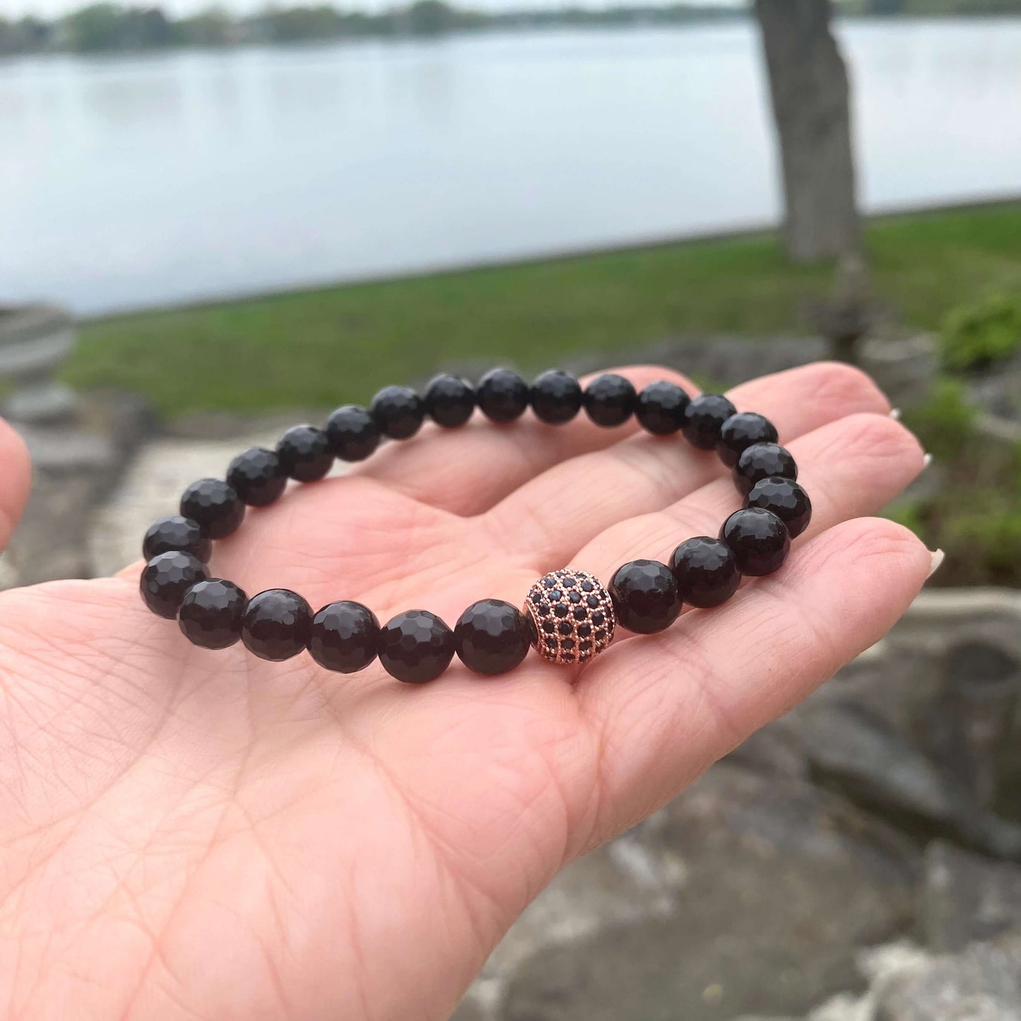 Black Onyx Mens Bracelet – Stretch Design with Rose Gold Accents displayed on a hand outdoors near a lake.