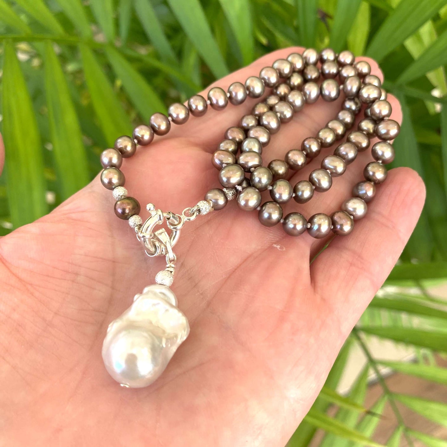 Hand holding bronze pearl necklace with removable white pendant against green leaves, showing iridescence and organic form.