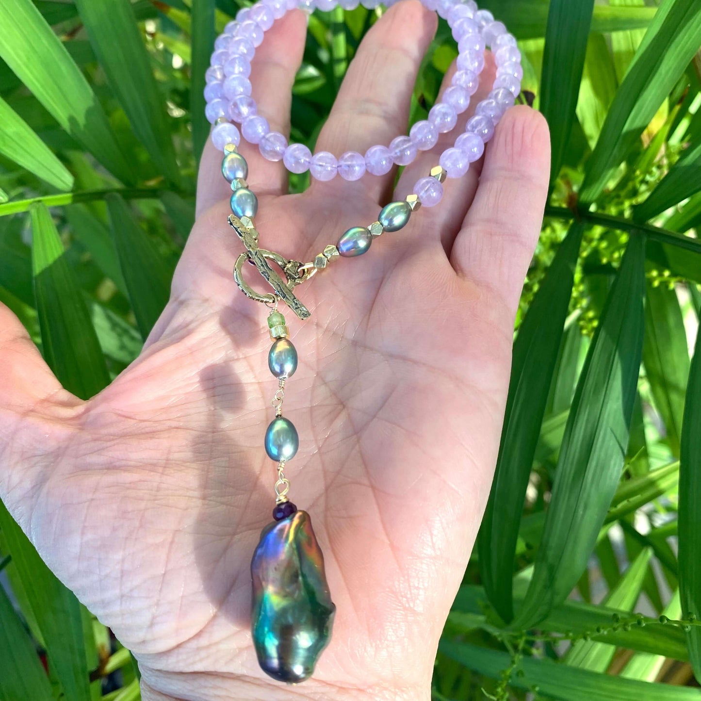 Amethyst Pearl Necklace with lavender beads and black baroque pearl held in hand against green foliage background.
