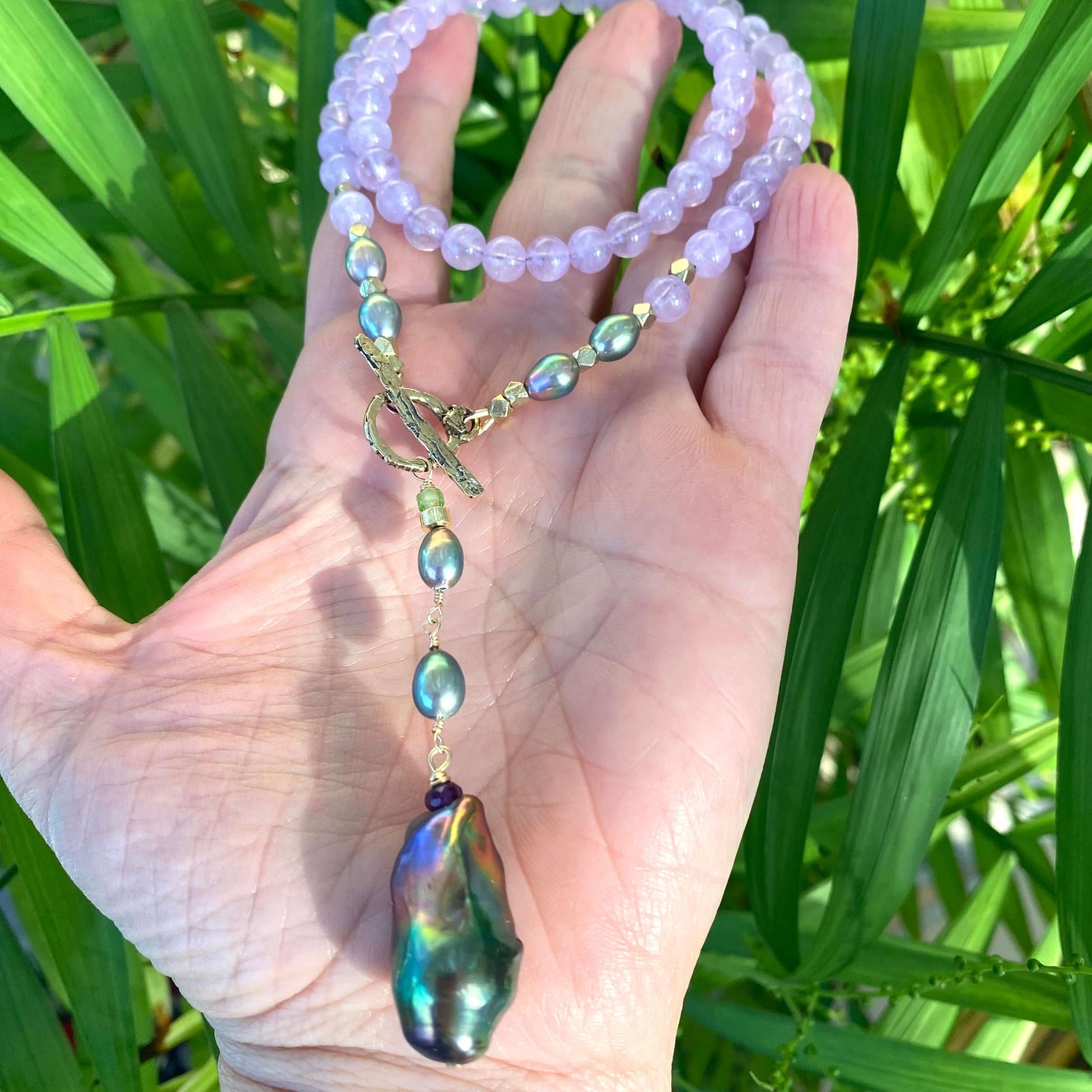 Amethyst Pearl Necklace with lavender beads and black baroque pearl held in hand against green foliage background.