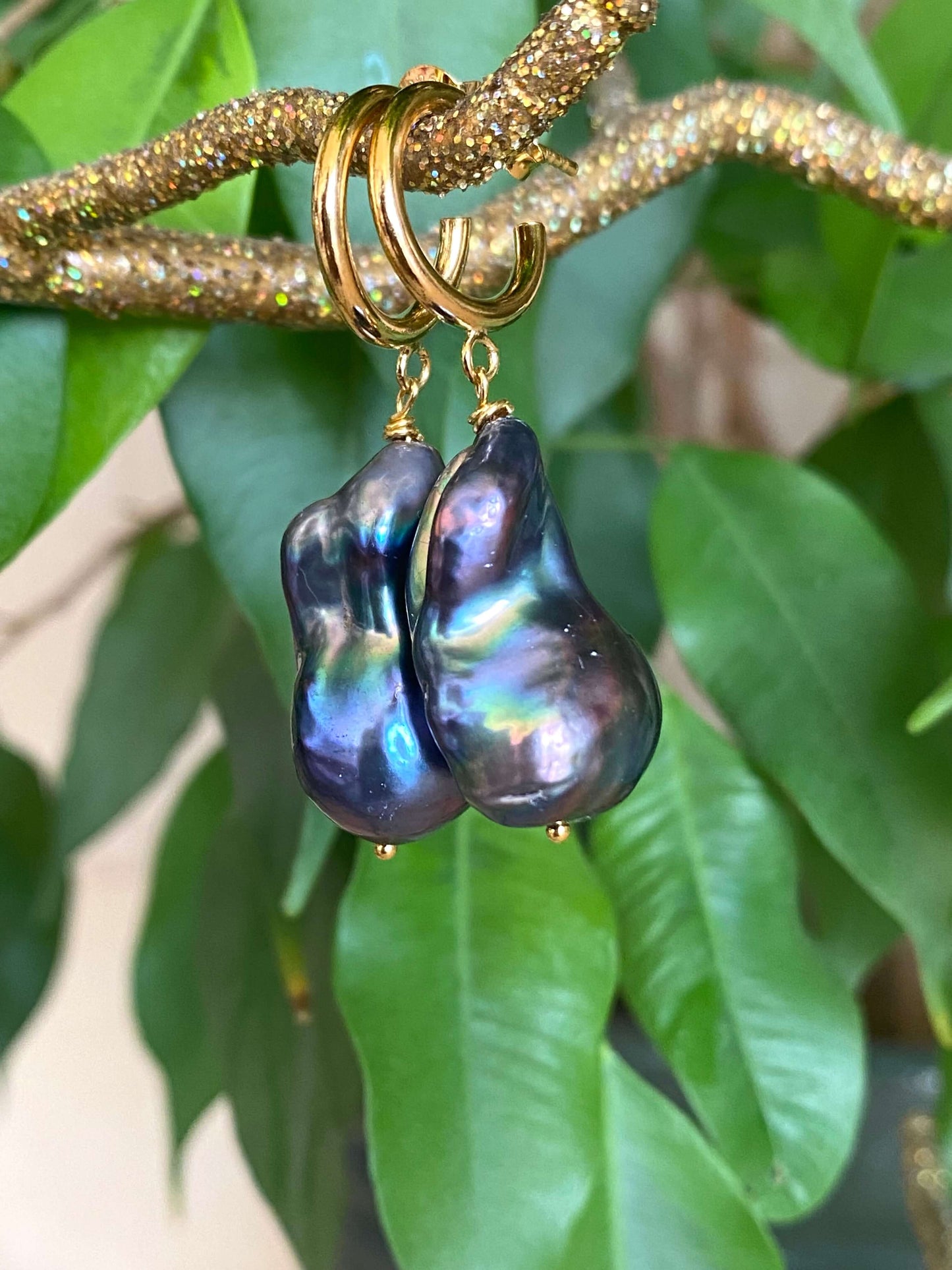 Baroque pearl hoop earrings in black peacock hues with gold vermeil hoops against green leaf background