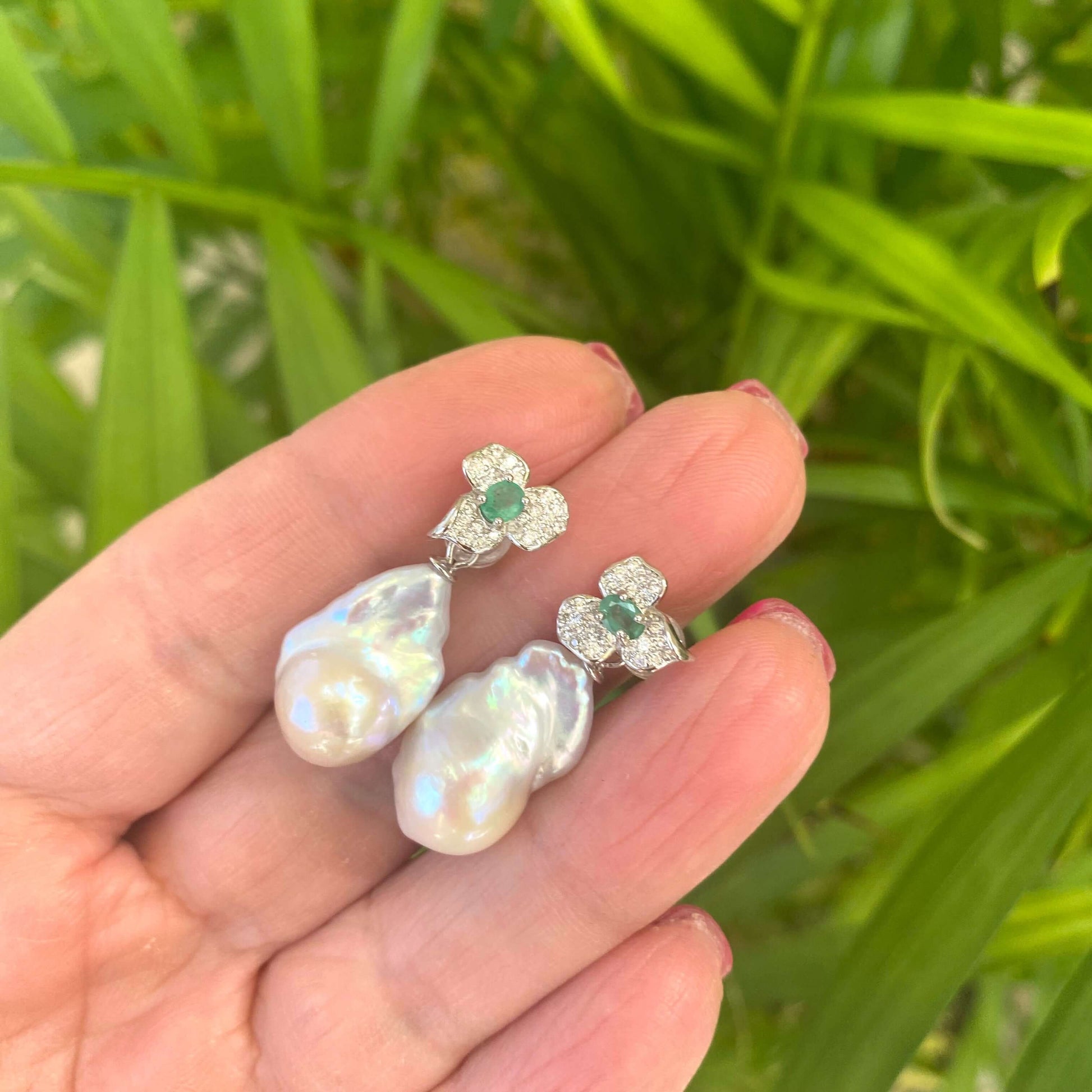 Emerald stud earrings with pearl drops held in hand against green leaf backdrop
