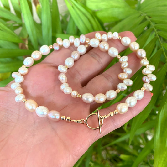 Pearl toggle necklace with pastel pearls and 14K gold filled clasp, held in hand against green backdrop.