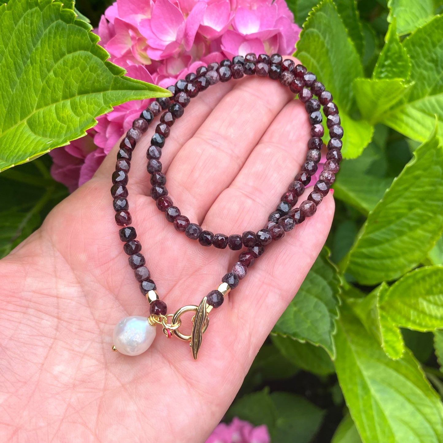 Garnet toggle necklace with baroque pearl charm displayed on hand among vibrant green leaves and pink flowers.