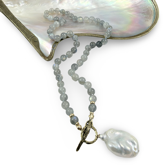 A beaded necklace with light gray baroque pearls and a large white baroque pearl pendant, featuring gold-toned hardware and a toggle clasp, displayed on a mother-of-pearl fan against a white background.
