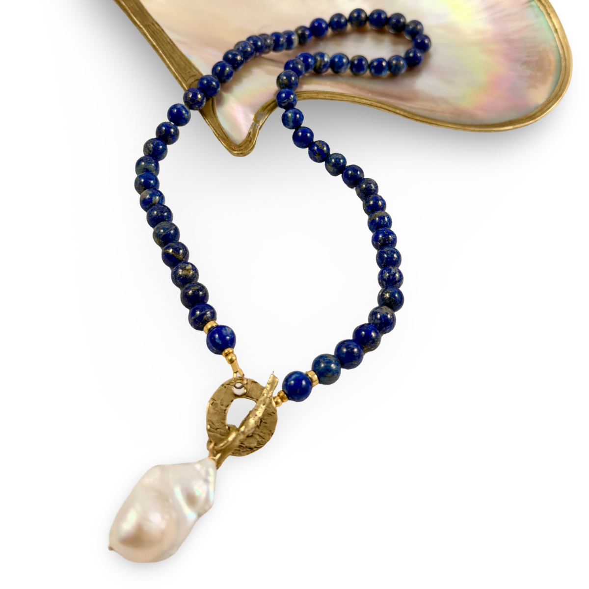 A beaded necklace with deep blue lapis lazuli stones, gold-toned hardware, and a large baroque pearl pendant, displayed on a white background with a mother-of-pearl shell prop.