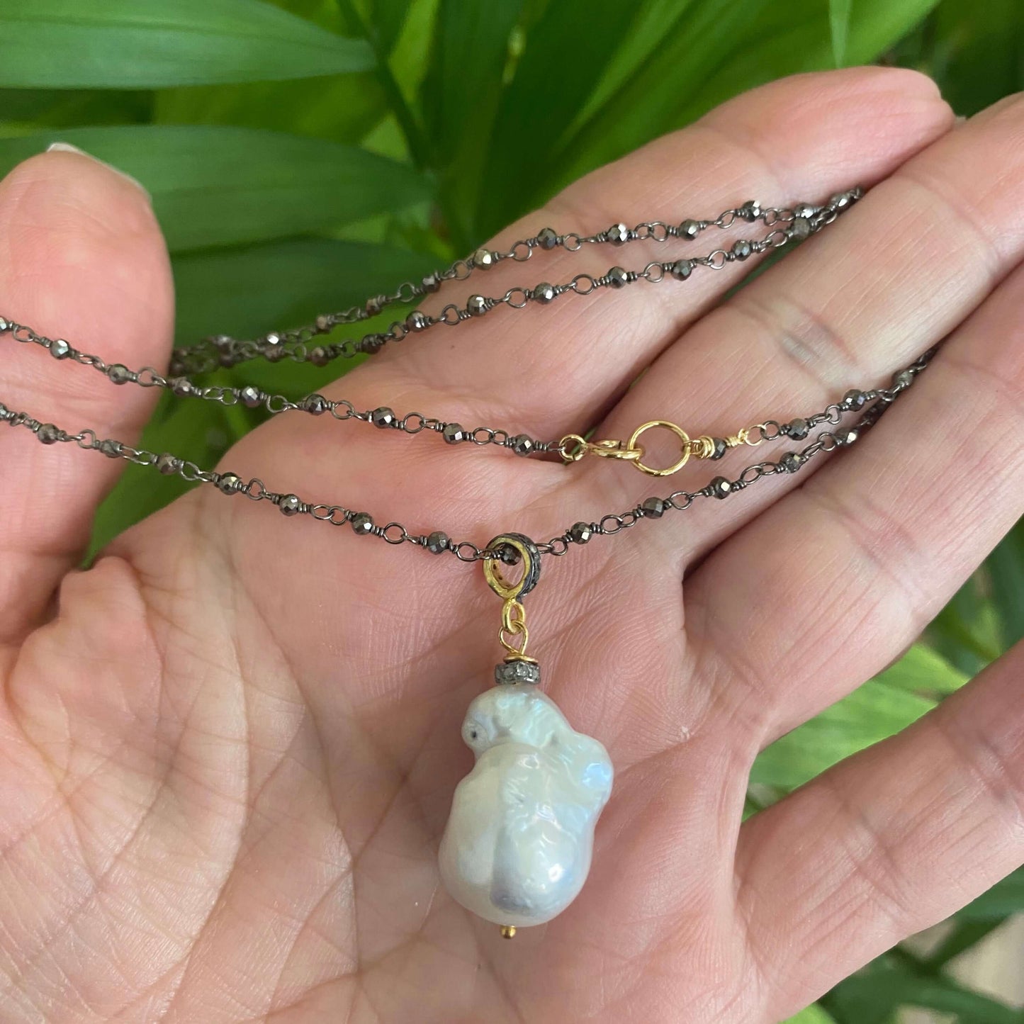Close-up of pyrite rosary chain necklace held near leaves highlighting baroque pearl and pavé detail