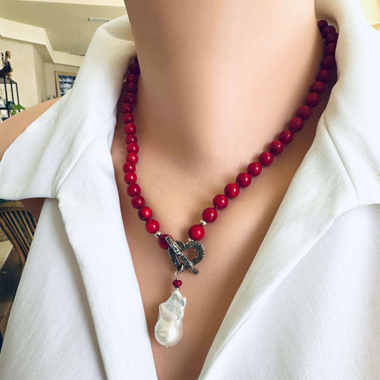 A close-up of a red beaded necklace with a silver-toned heart-shaped clasp and a white baroque pearl pendant, worn on a mannequin or model with a white collared shirt.
