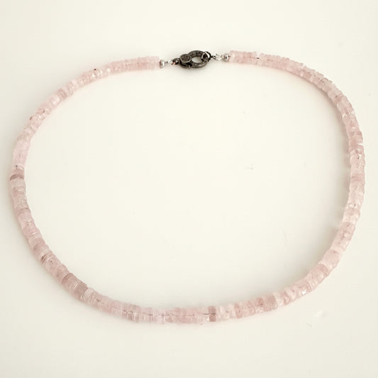 A delicate necklace made of small, faceted light pink beads, featuring a dark metal clasp, displayed on a plain off-white background.
