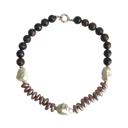 A beaded necklace featuring dark brown faceted beads, two white baroque pearls, and a row of iridescent mauve-pink spiral baroque pearls, finished with a silver clasp on a light beige background.