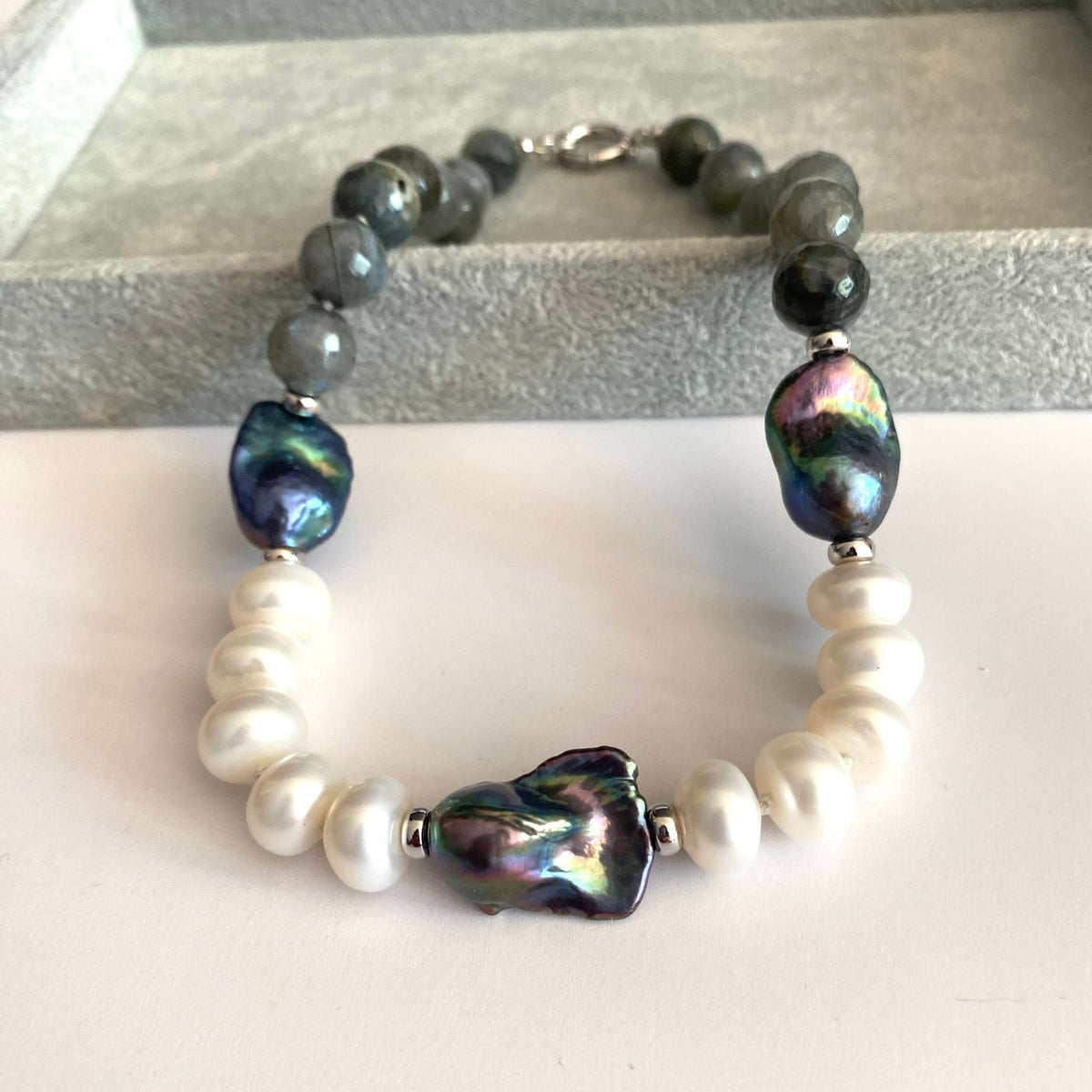 A beaded necklace featuring white baroque pearls, dark gray beads, and three large iridescent green-blue baroque shell beads, displayed on a light surface.