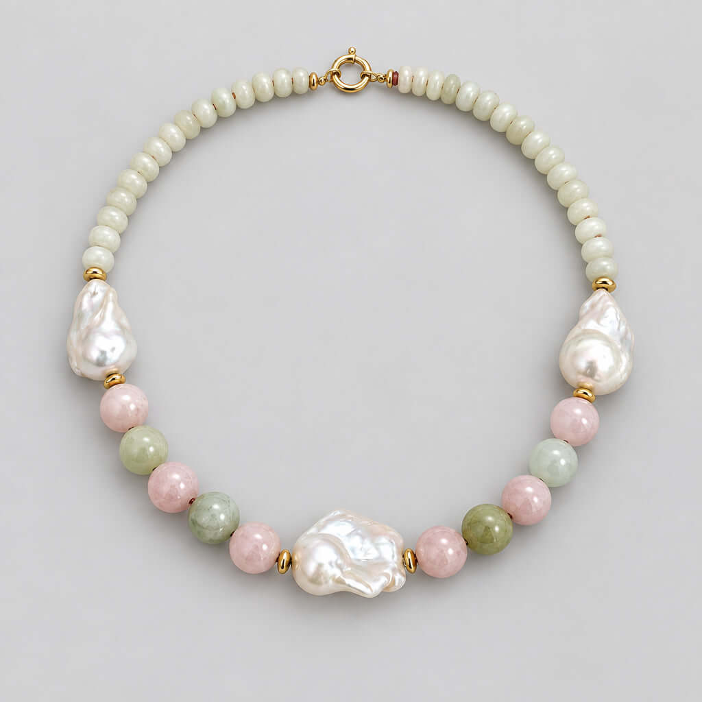 A handmade beaded necklace featuring a strand of small cream-colored beads, irregular white baroque pearls, polished pink and green beads, and gold spacer rings, displayed on a neutral light beige background.