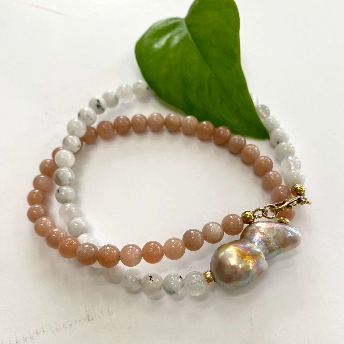 A double-strand beaded bracelet with peach and white beads, featuring a large iridescent baroque pearl and gold-tone hardware, displayed on a white surface with a green leaf.