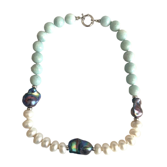 A beaded necklace featuring light mint green round beads, white baroque pearls, and three iridescent blue-purple abalone shell beads, arranged in a circle on a light beige background.