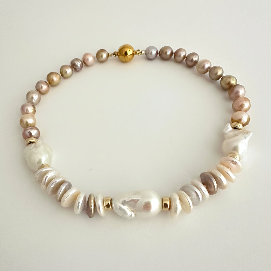 A beaded necklace featuring a mix of iridescent baroque pearls in beige, brown, and white, accented with small gold spacer beads and a gold clasp, displayed on a neutral light gray background.