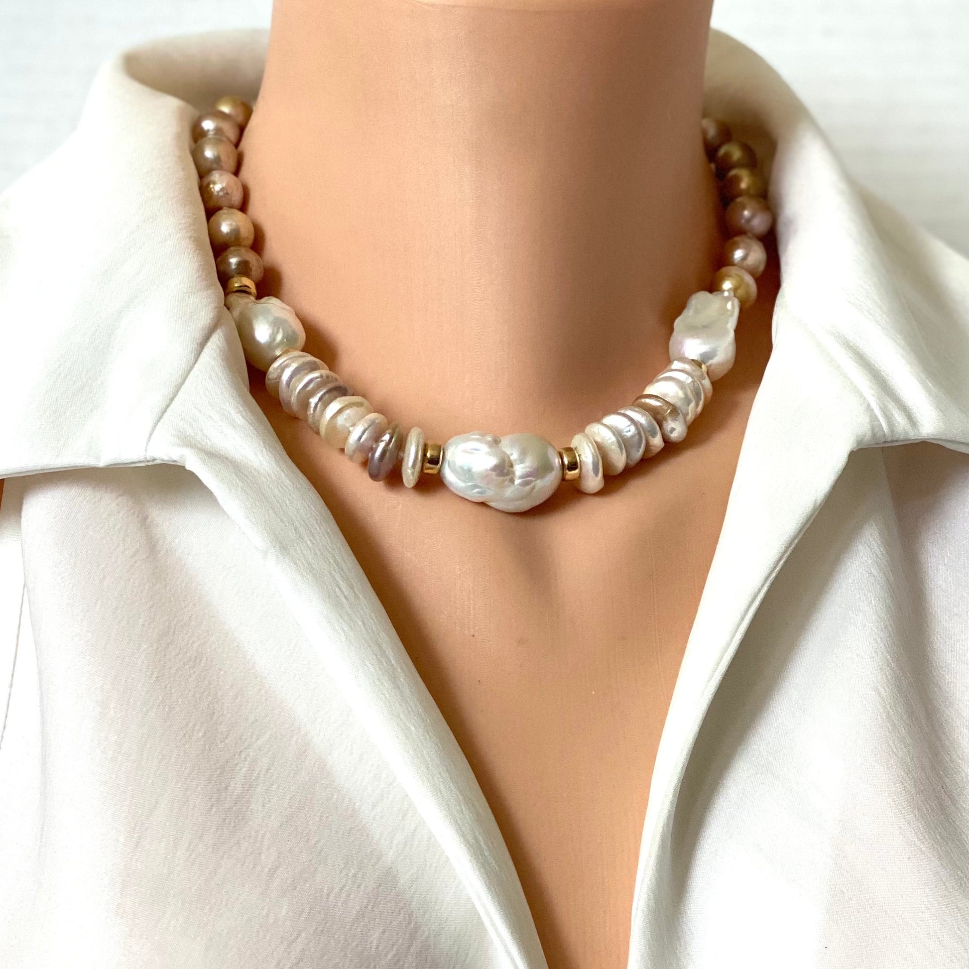A pearl necklace with mixed white, cream, and beige baroque pearls and small metallic spacer beads, shown on a mannequin torso wearing a white satin shirt.