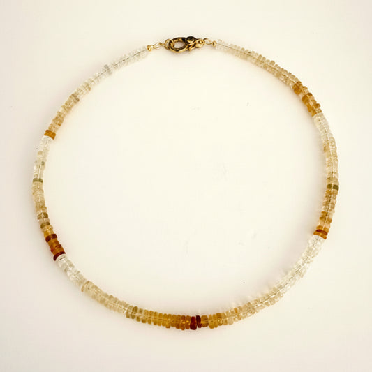 A beaded necklace made of small translucent and opaque beads in white, clear, and warm amber-brown tones, with a dark metal clasp, laid flat on a light off-white background.