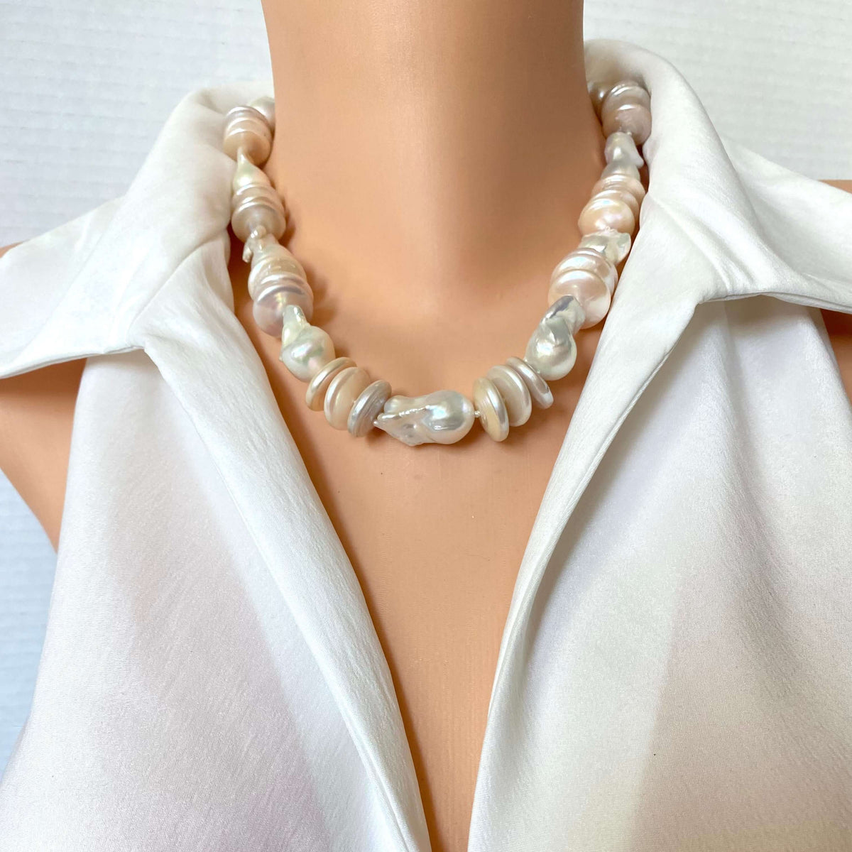 A close-up of a pearl necklace with irregular baroque-style pearls in white, cream, and light pink tones, worn on a mannequin torso wearing a white satin blouse.