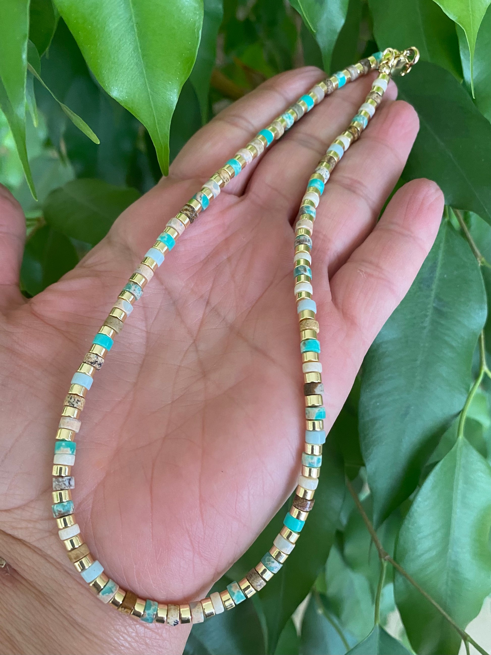 Gemstone choker necklace with multi color beads and hematite held in a hand surrounded by green leaves.