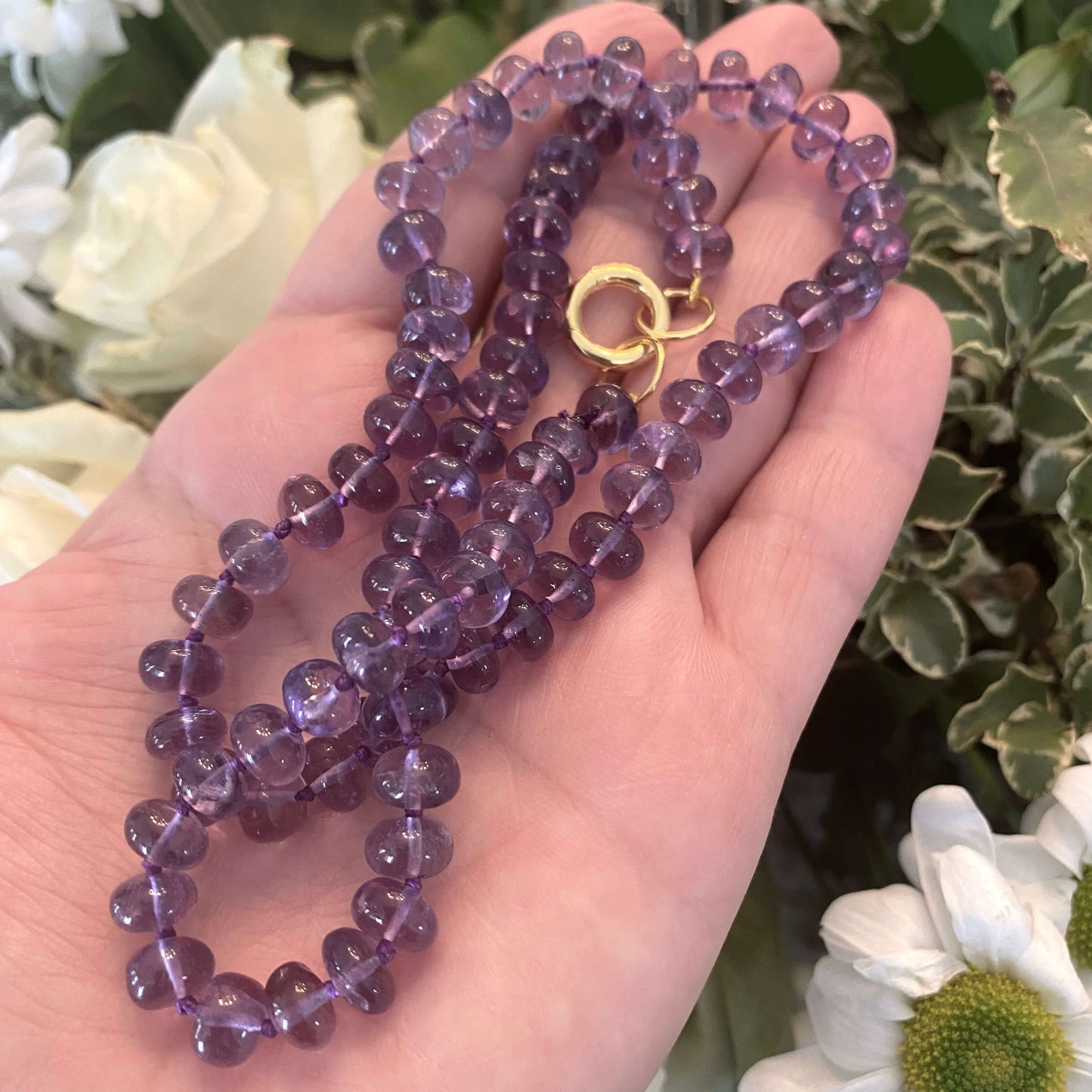 Amethyst candy necklace featuring Brazilian amethyst beads in rich purple tones, showcased in a hand against floral backdrop.